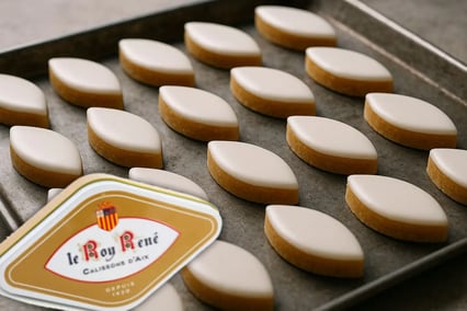 Une boîte de calissons d'Aix de la marque Roy René est présentée en premier plan. A l'arrière, plusieurs rangées de calissons sont déposées sur un plateau.