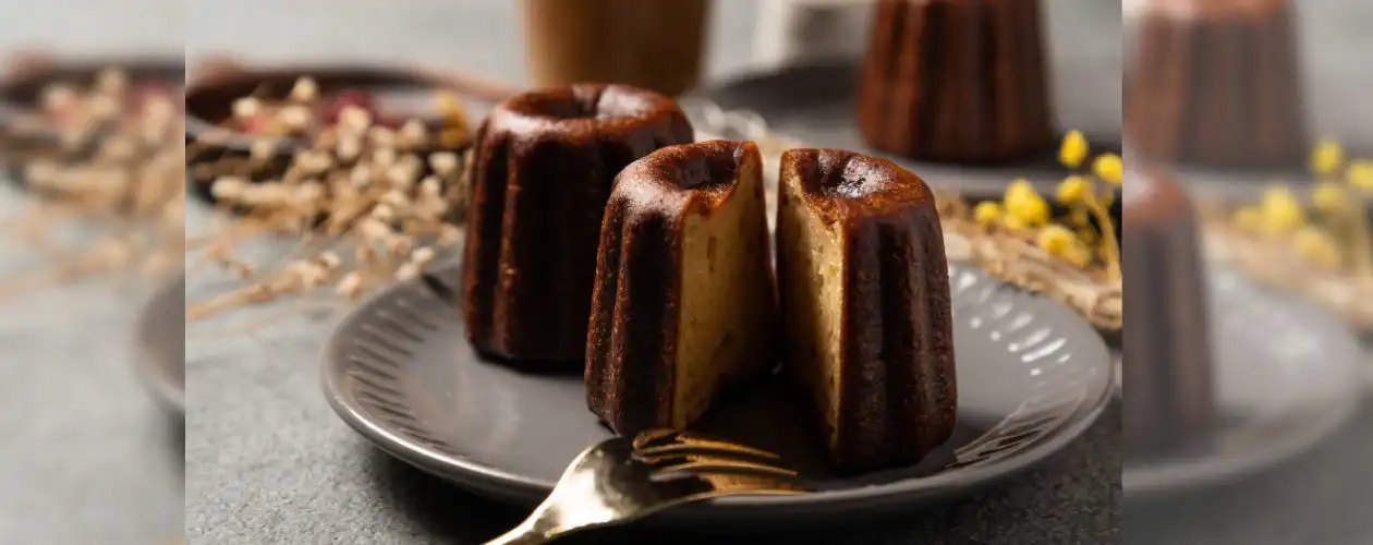 Deux cannelés posés dans une assiette ; l'un des cannelés est coupé en deux laissant apparaître son cœur moelleux dans la croûte caramélisée